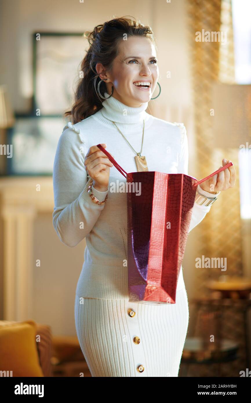 Lächelnde, trendige Hausfrau in weißem Pullover und Rock mit roter Einkaufstasche im modernen Wohnzimmer am sonnigen Wintertag. Stockfoto