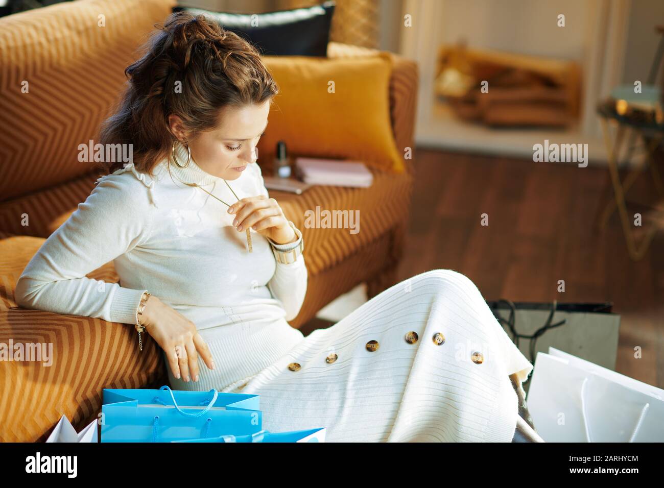 Besorgt junge Frau in weißem Pullover und Rock in der Nähe von Divan mit Einkaufstaschen im modernen Zuhause am sonnigen Wintertag. Stockfoto