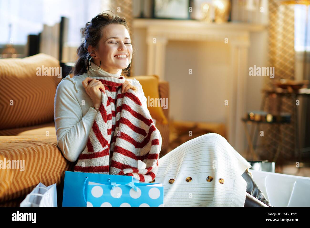Lächelnde trendige Frau in weißem Pullover und Rock in der Nähe eines Sofas mit Einkaufstaschen und einem Pullover im modernen Haus am sonnigen Wintertag. Stockfoto