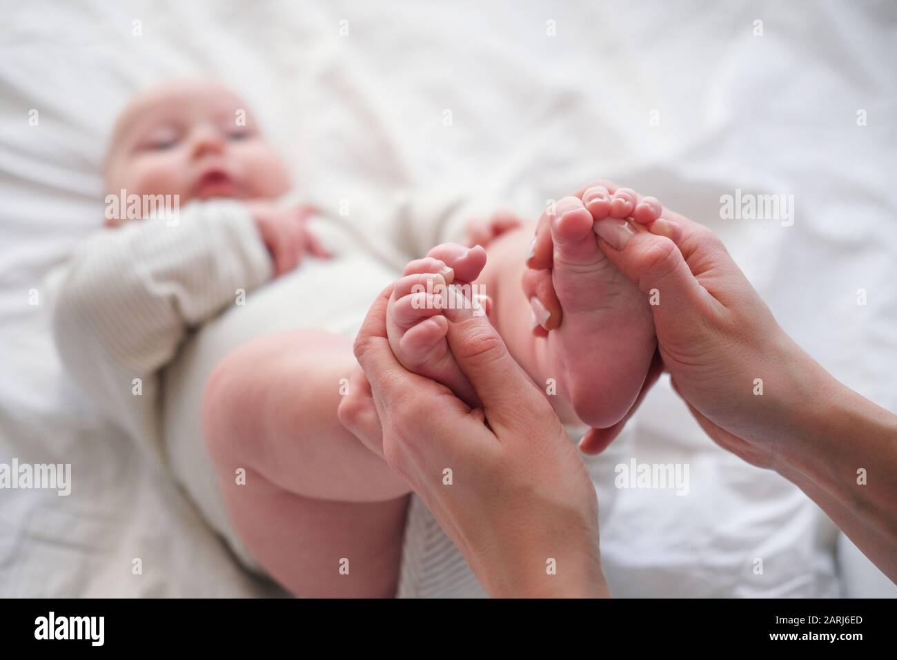 Baby feets in den Händen der Mutter. Die junge kaukasische Frau massiert zu Hause für glückliches Säuglingsbaby auf weißem Bett. Babypflege, Sport und glückliche Mutterschaft. Stockfoto
