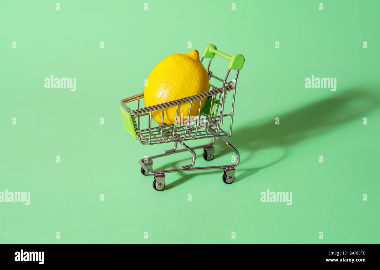 Einzelne Zitrone in einem Supermarktwagen auf grünem Grund. Mini-Einkaufswagen mit nur einer Zitrone darin. Konzept des Lebensmitteleinkaufs. Kleines Budgetkonzept Stockfoto
