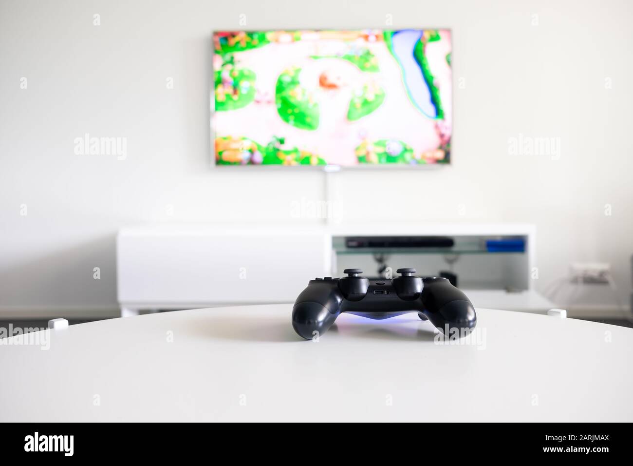 Eine Ansicht eines Logo-freien, schwarzen Videobame-Controllers auf einem weißen Tisch. Der Fernseher befindet sich im Hintergrund. Stockfoto