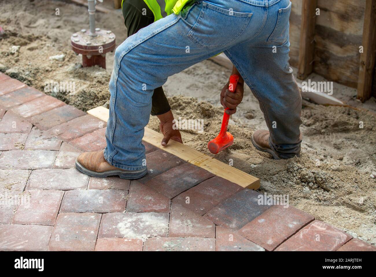 Backstein-Schicht, die seinen orangefarbenen Hammer oder Hammer gegen einen 2x4 schlägt, um die frisch verlegten Fertiger auf dem Gehweg einzustellen. Stockfoto