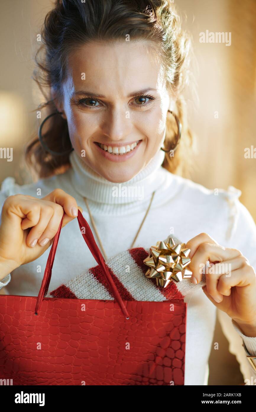 Porträt einer fröhlichen jungen Frau in weißem Pullover und Rock mit roter Einkaufstasche, die im modernen Haus im sonnigen Wintertag gekauften Pullover auszieht. Stockfoto