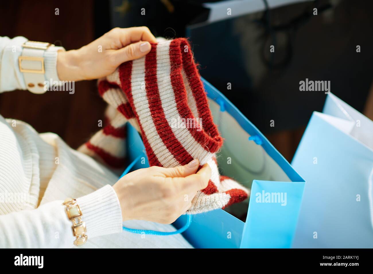 Nahaufnahme von stilvoller Frau in weißem Pullover und Rock mit Einkaufstaschen Überprüfung gekaufter Pullover im modernen Haus am sonnigen Wintertag. Stockfoto