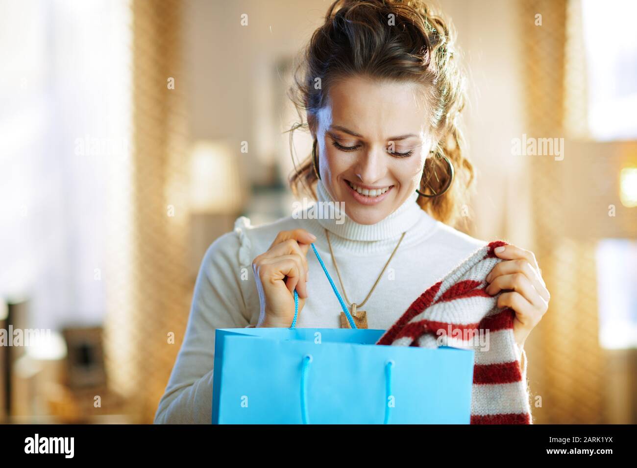 Fröhliche, trendige Hausfrau in weißem Pullover und Rock mit blauer Einkaufstasche, die im modernen Haus im sonnigen Wintertag einen Pullover gekauft hat. Stockfoto