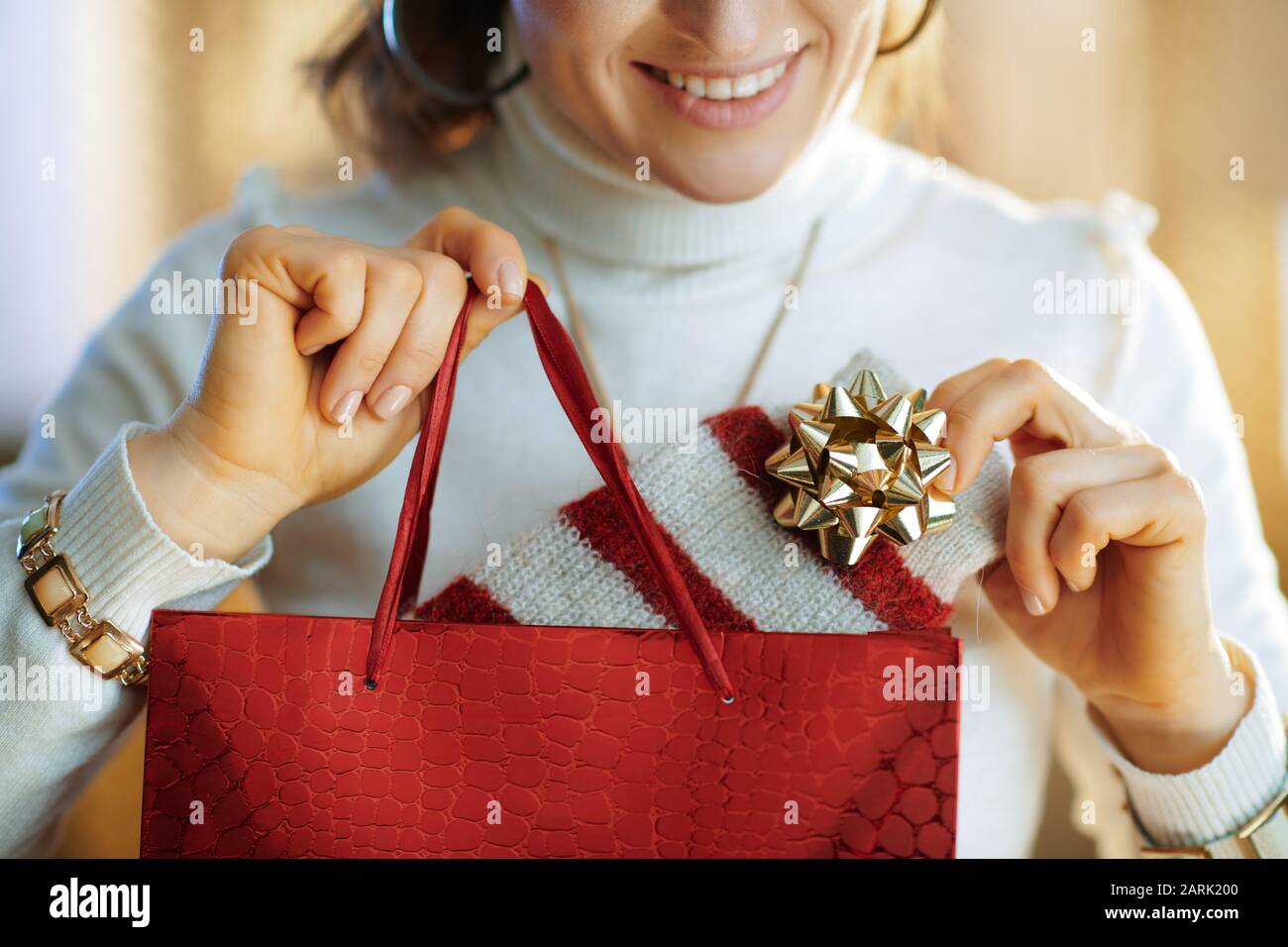 Die elegante 40 Jahre alte Hausfrau im weißen Pullover und Rock mit roter Einkaufstasche, die im sonnigen Winter einen Pullover im modernen Zuhause auszieht Stockfoto