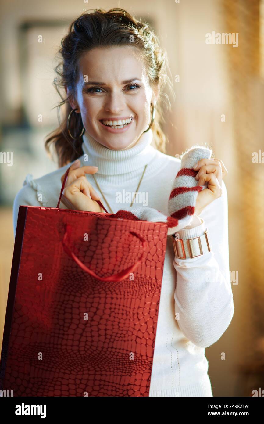 Porträt einer fröhlichen, stilvollen Frau in weißem Pullover und Rock mit roter Einkaufstasche, die den gekauften Pullover im modernen Zuhause am sonnigen Wintertag auszieht. Stockfoto