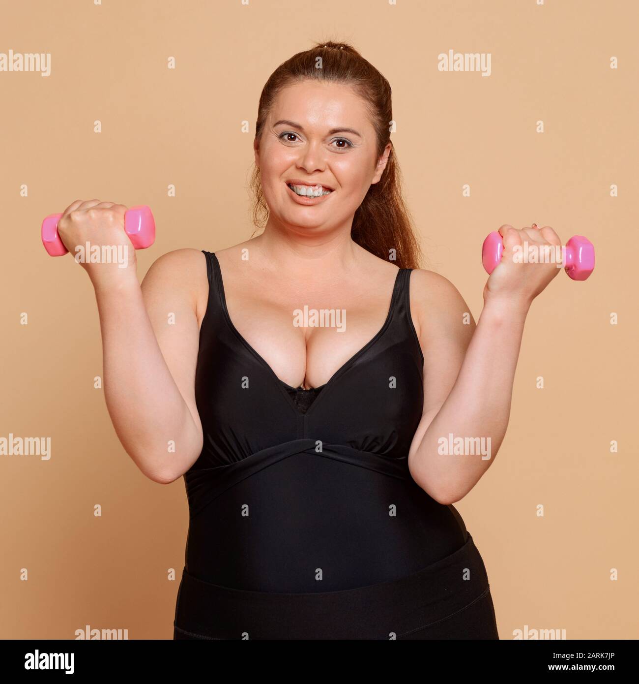 Positiv übergewichtige Frau mit Hanteln auf beigem Hintergrund Stockfoto