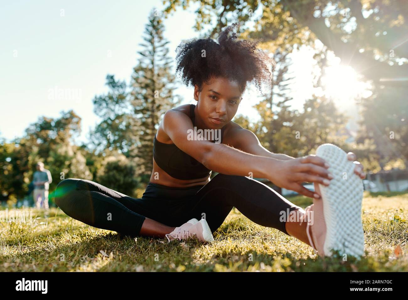 Portrait einer entschlossenen afroamerikanischen jungen Frau, die morgens auf grünem Gras sitzt, Sonnenlicht, die ihre Beine mit Blick auf die Kamera streckt Stockfoto