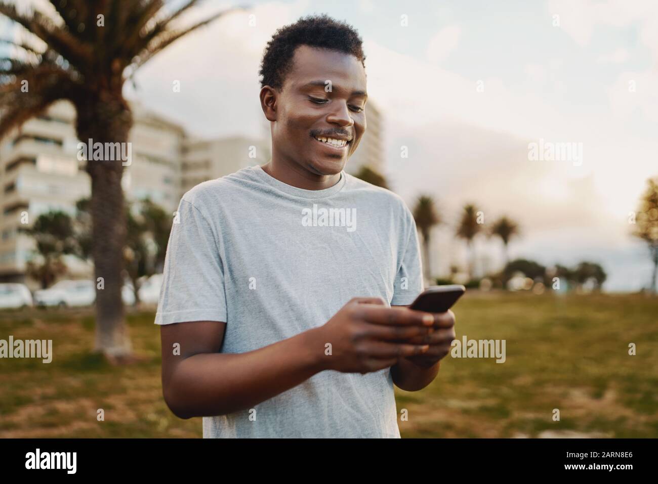 Portrait eines sportlichen jungen afroamerikanischen Mannes, der lächelt, während er Nachrichten auf dem Handy im Park textet Stockfoto