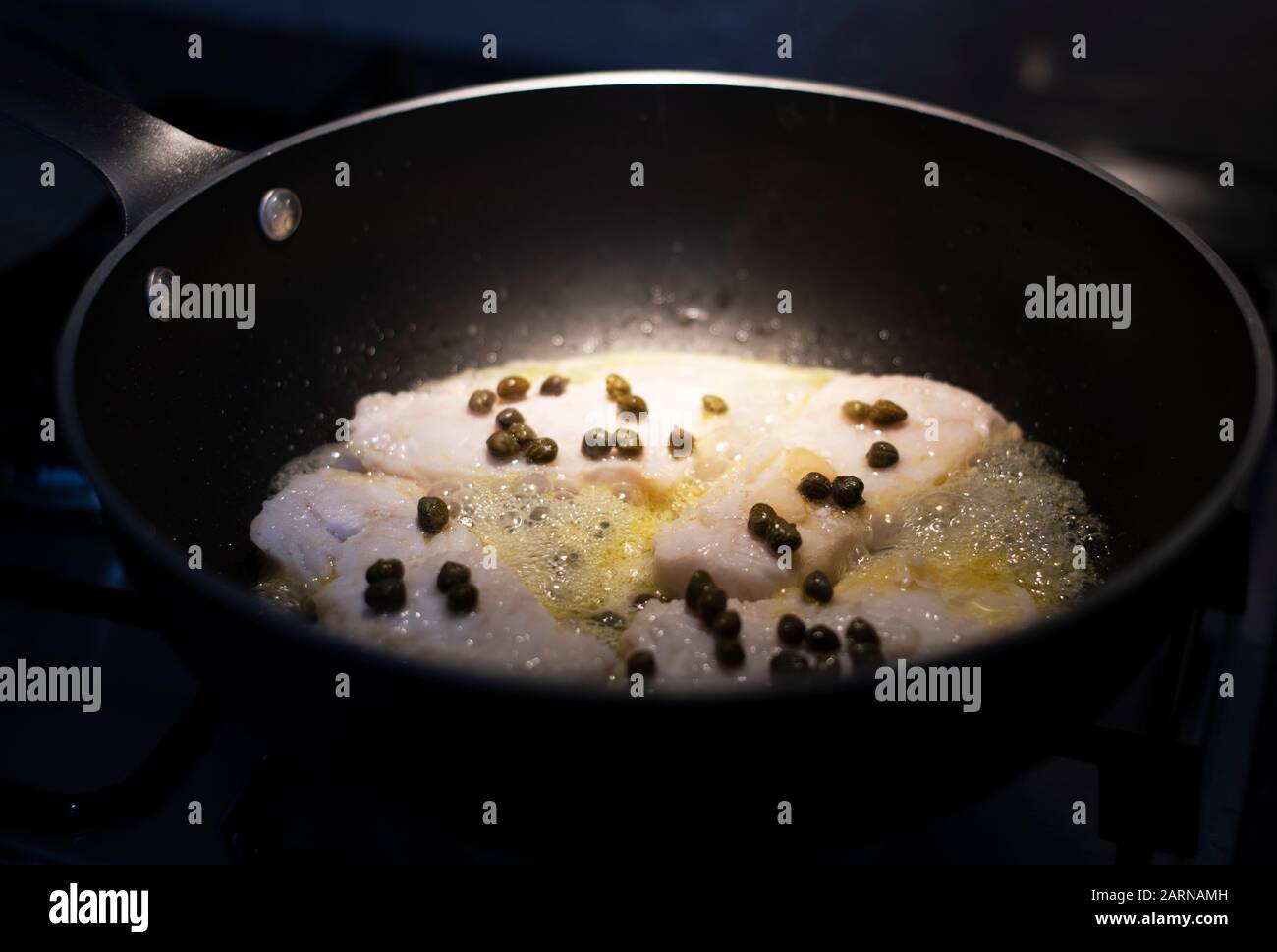 Fischfilets, die in heißem Öl und Wein mit Kapern in einer Bratpfanne über einem Gasbrenner in Nahaufnahme mit Beleuchtung des Essens sizzeln Stockfoto