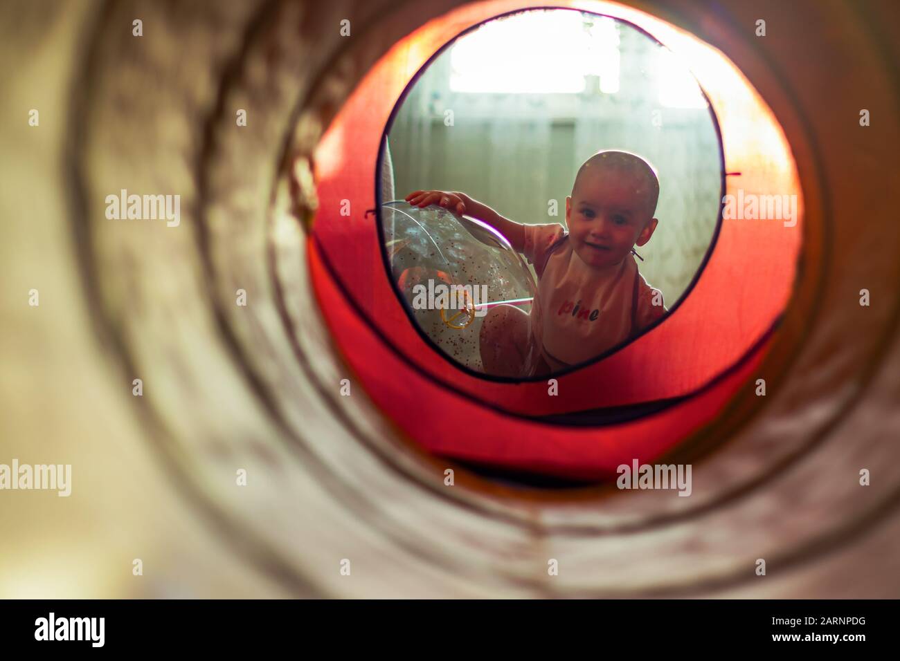 Ein süßes kleines Mädchen, das sich an den transparenten Luftballon lehnt und durch einen bunten Tunnel auf die Kamera blickt. Stockfoto