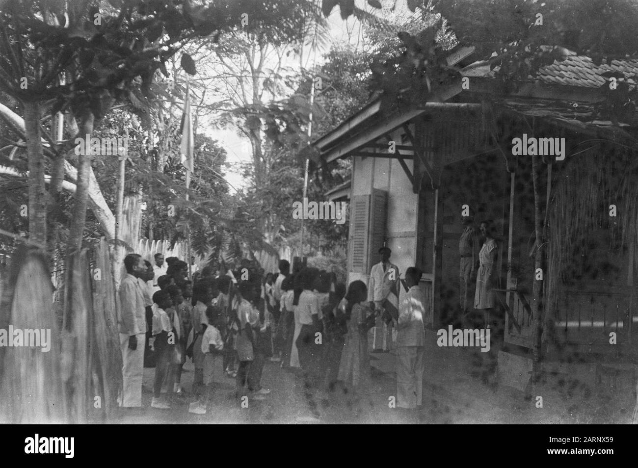 Neuguinea-Serie [eine Gruppe von Kindern steht mit holländischen Flaggen für dekorierte Bögen. Auf der Vorhalle eines Hauses sind ein niederländischer Mann in Uniform und eine Holländerin. Dafür ein indigener Lehrer?] Datum: Oktober 1948 Ort: Neuguinea Stockfoto