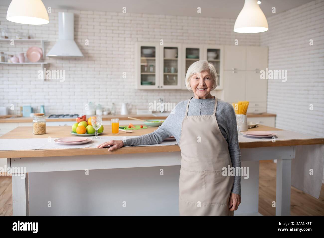 Weißhaarige Frau in Schürze, die vor dem Küchentisch steht. Stockfoto