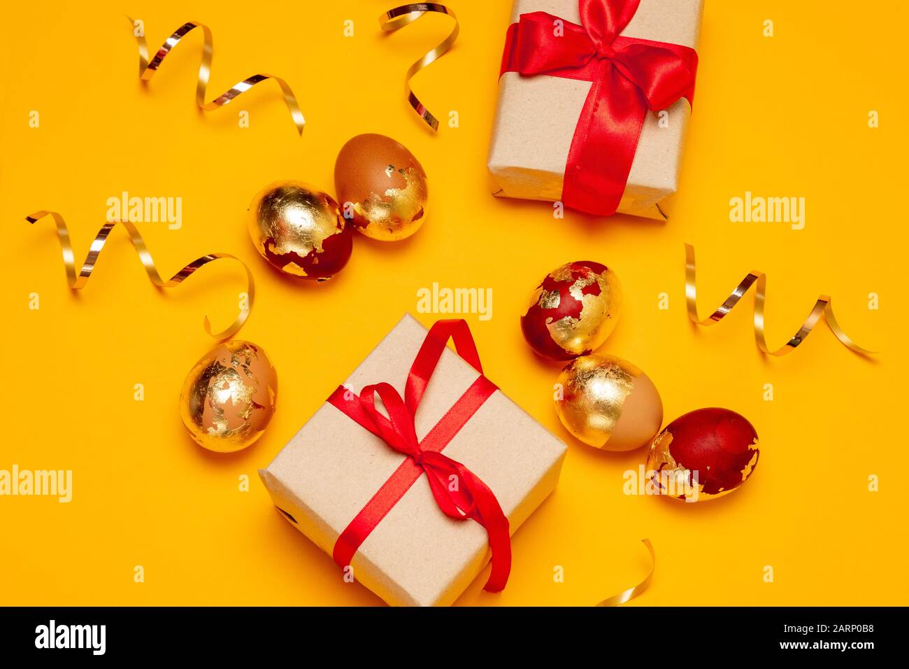 Überraschungsgeschenkkasten in Handwerkspapier und mit rotem Bogen und goldenen Eiern auf gelbem Hintergrund. Glitzernder Serpentin. Osterfeier Stockfoto