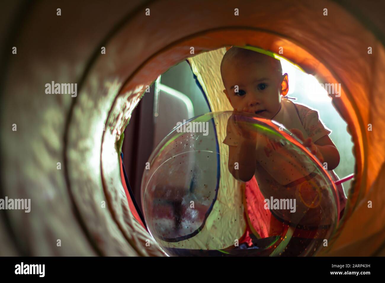 Ein süßes kleines Mädchen, das sich an den transparenten Luftballon lehnt und durch einen bunten Tunnel auf die Kamera blickt. Stockfoto