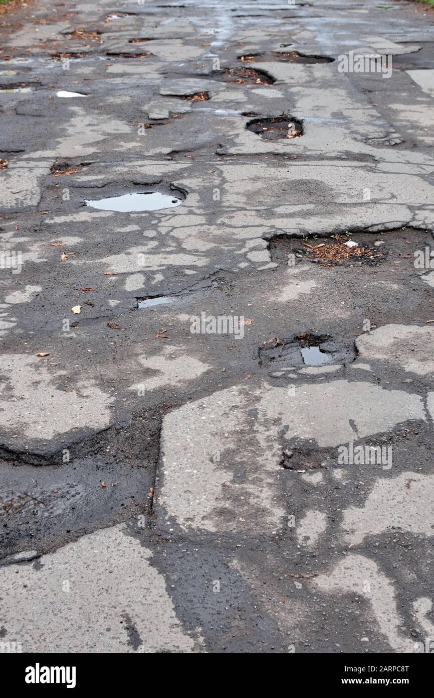 Alte zerstörte Asphaltstraße mit Schlaglöchern und Erschütterungen. Nasse Herbststraße mit Blättern. Stockfoto