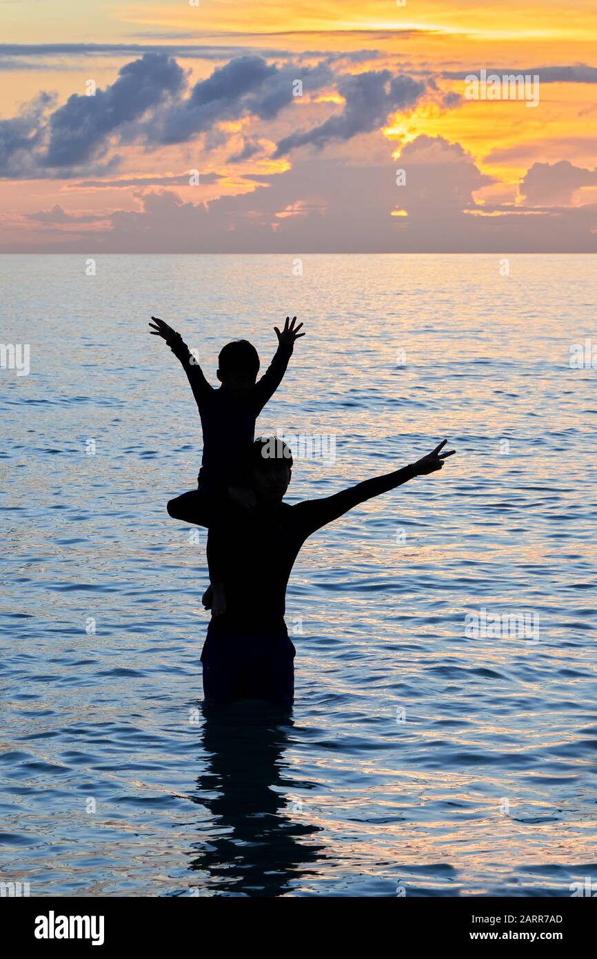 Silhouette eines Vaters, der seinen jungen Jungen auf den Schultern hält und im Meer entlang des White Beach auf Boracay Island in farbenfrohem Sonnenuntergang steht Stockfoto