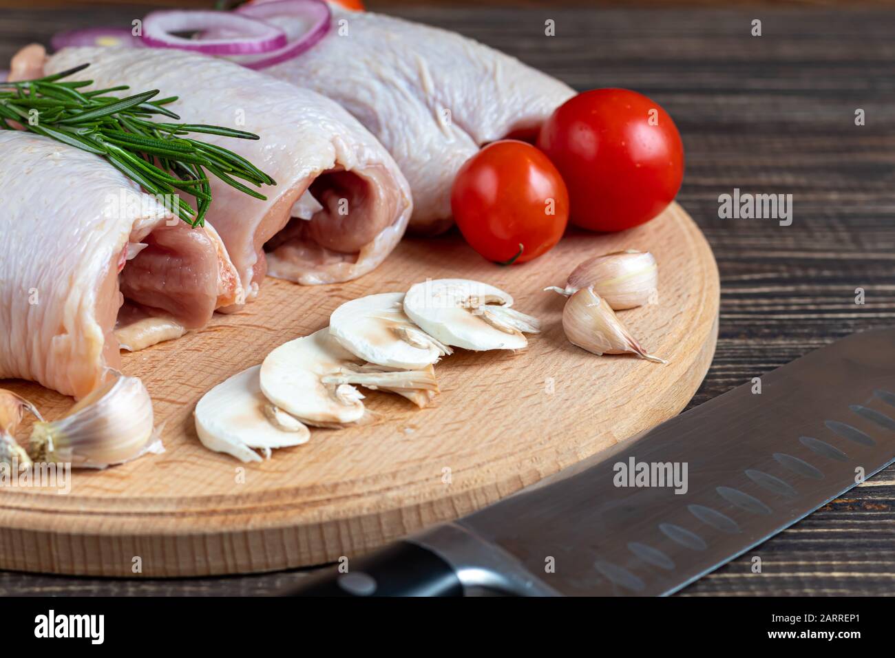 Auf einem hellen Holzbrett, frische, häutige Hühnerschenkel, neben Pilzen, mit Tomaten, roten Zwiebeln und Rosmarin. Rezept des Gerichts. Zutaten. Kopieren Stockfoto