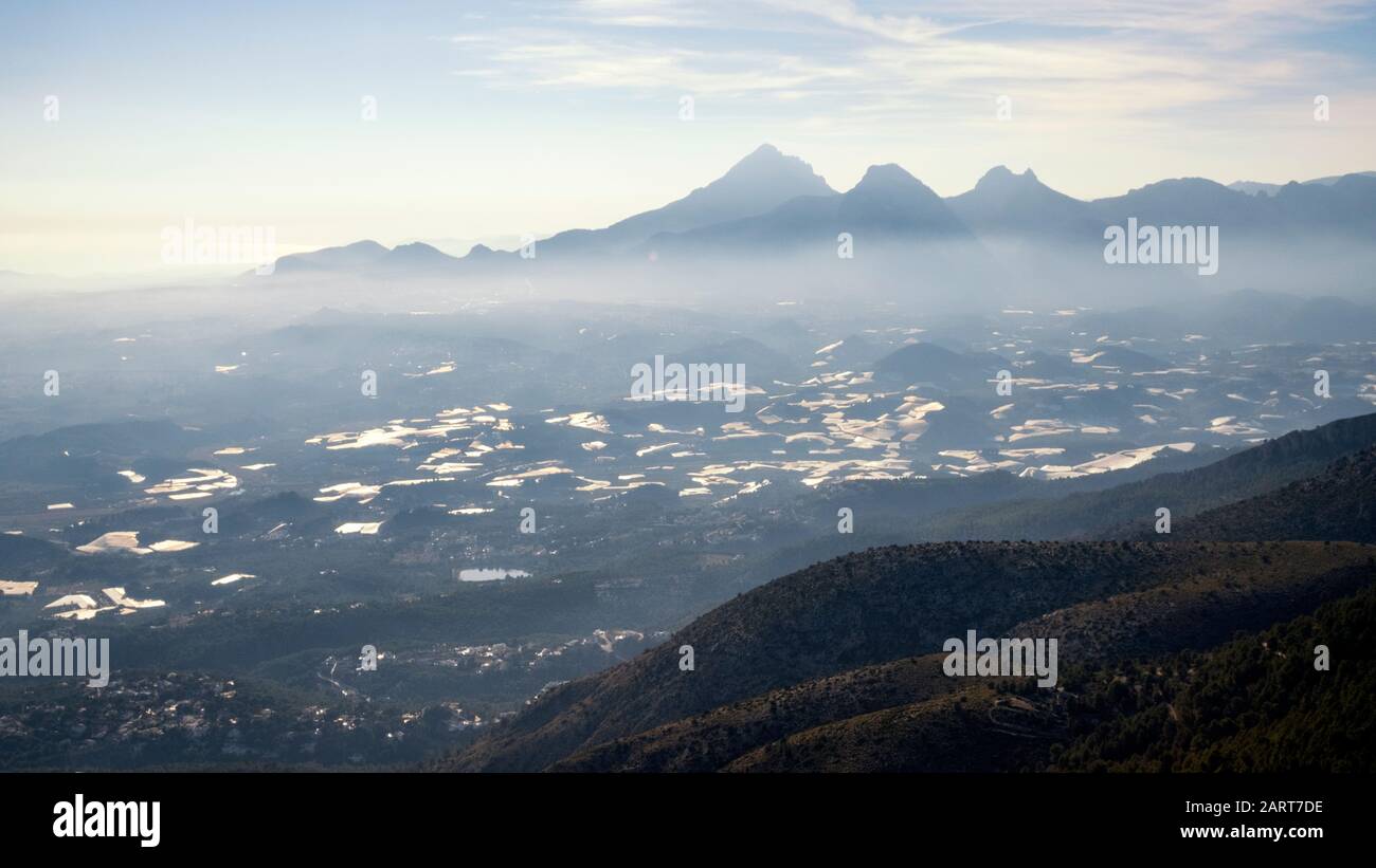 Berge der Sierra de Aitana und Gewächshäuser zu seinen Füßen in einem Dunstbereich von der Sierra de Bernia (Marina Baixa, Costa Blanca, Alicante, Spanien) Stockfoto