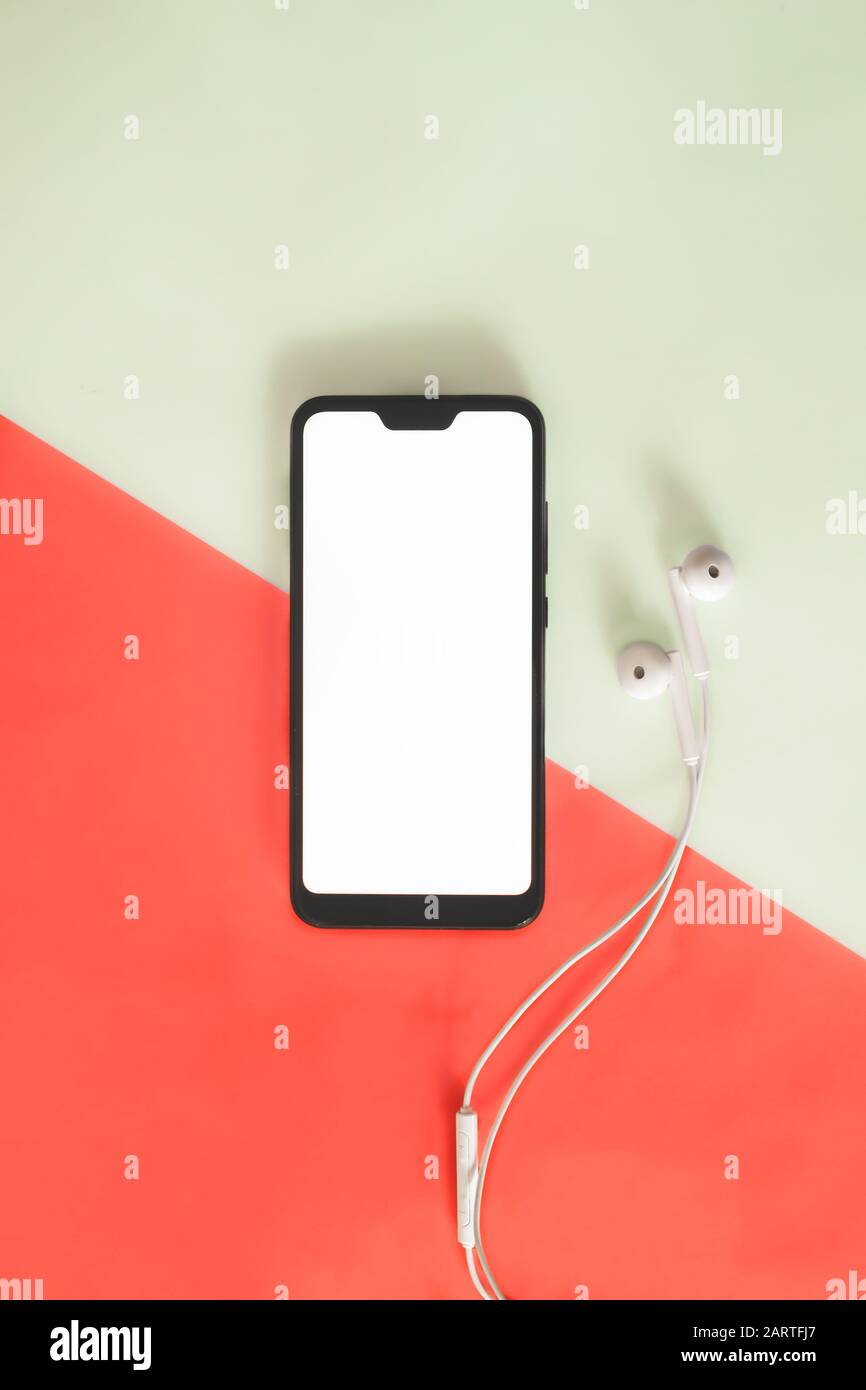 Modernes Smartphone mit weißem Bildschirm und Kopfhörern auf lebendigem Hintergrund. Draufsicht eines Telefons und Headsets vor rotem und grünem Hintergrund Stockfoto