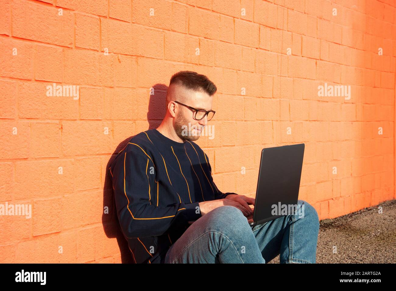 Jungen kaukasischen Mann in casual-wear mit überkreuzten Beinen auf dem Boden und Arbeiten am Laptop. Freelancer arbeiten außerhalb der Texteingabe auf Laptop Computer. Rote Wal Stockfoto