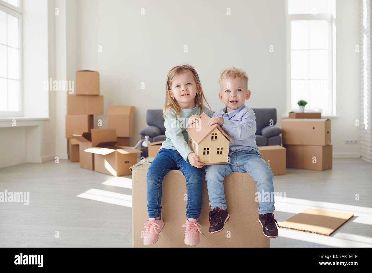 Zwei Kinder sitzen auf einer Schachtel, um sie zu bewegen und halten ein Modell eines Hauses in einem neuen hellen Raum. Stockfoto