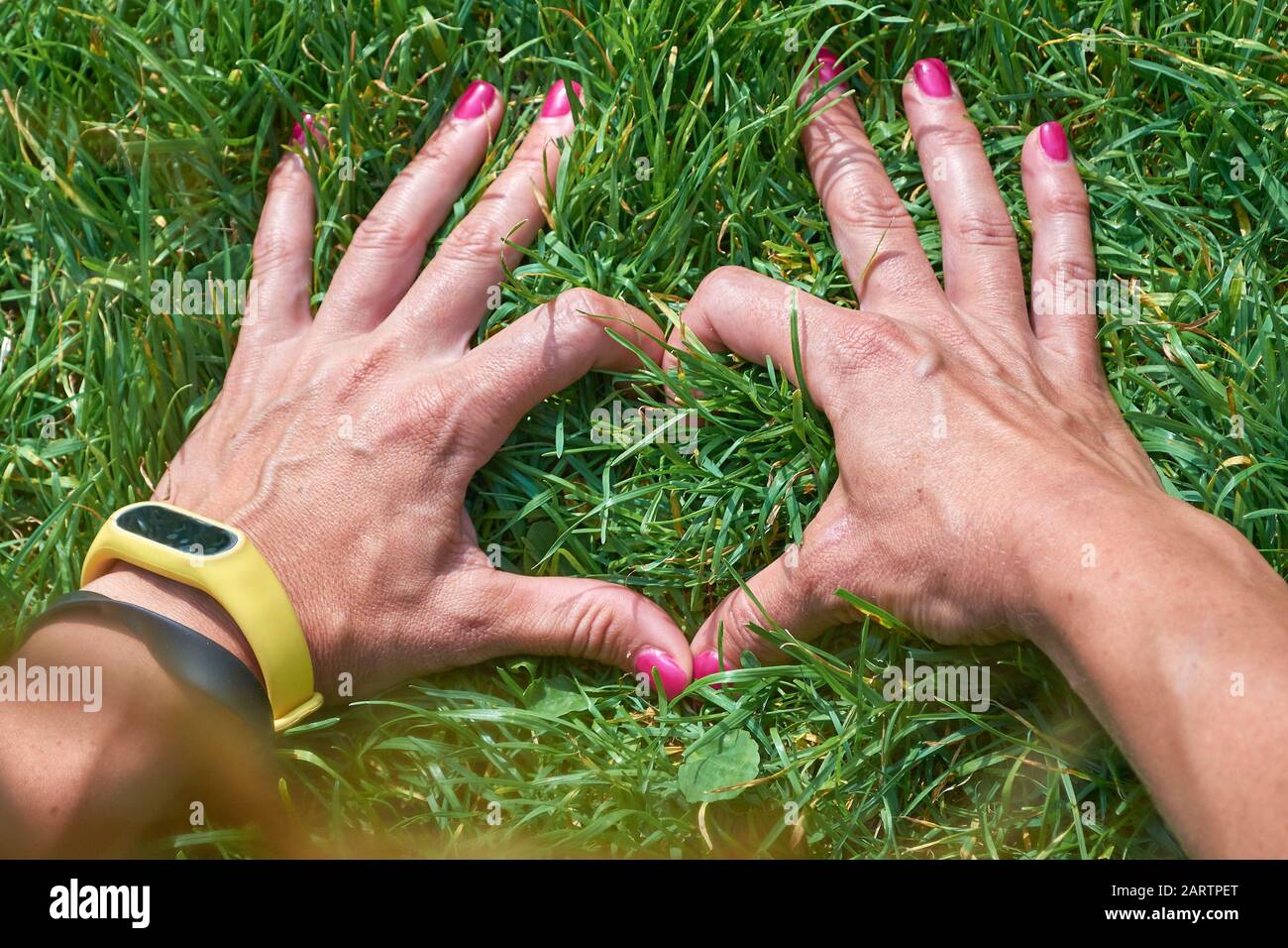 Frau, die mit ihren Händen ein herzförmiges Schild in perfektem grünem Gras trägt und dabei ein modernes Tech-Armband trägt. Verbindung zum Naturkonzept. Stockfoto