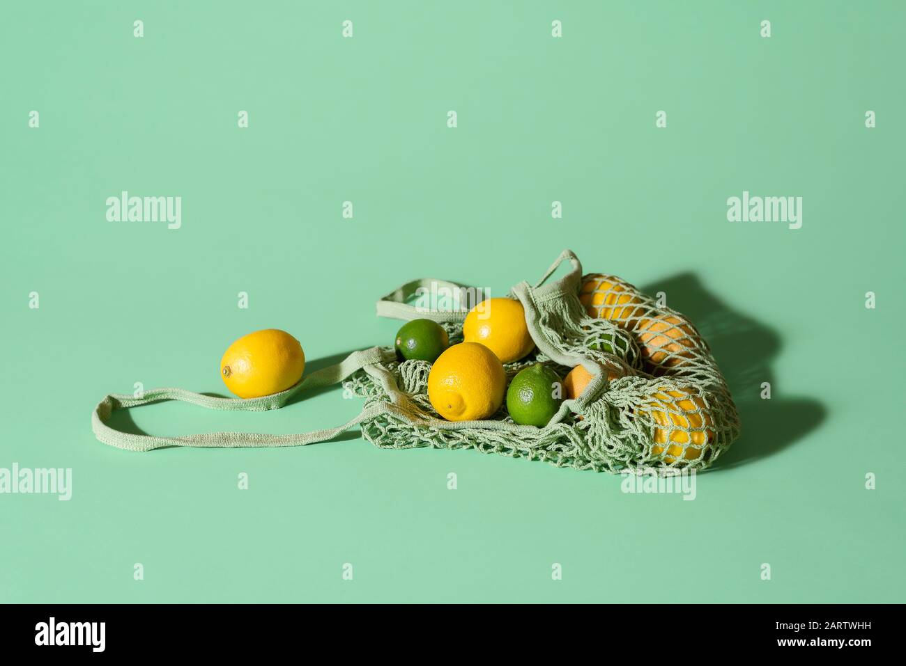 Zitronen und Limonen in einer wiederverwendbaren Einkaufstasche aus Stoff auf grünem Hintergrund. Umweltfreundliche Netztasche mit Zitrusfrüchten. Sommerfrüchte. Lebensmittel-Shopping-Konzept Stockfoto