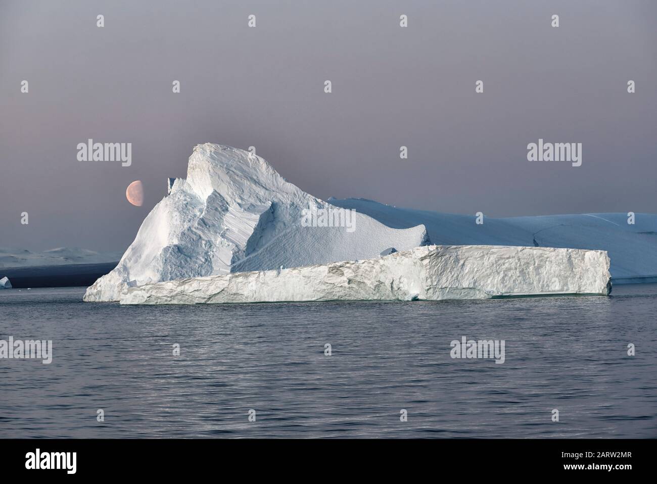 Riesiger schwimmender Eisberg in einem Fjord in der Dämmerung mit Mond auf der linken Seite. Scoresby Sund, Kangertittivaq, Grönland, Dänemark Stockfoto