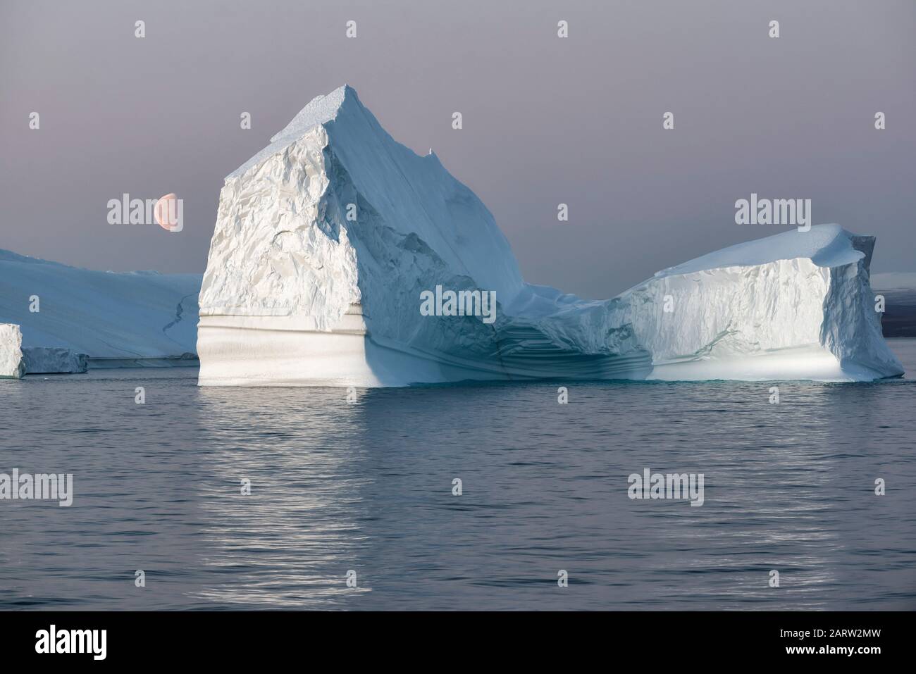 Riesiger schwimmender Eisberg in einem Fjord bei Sonnenuntergang mit Mond auf der linken Seite. Scoresby Sund, Kangertittivaq, Grönland, Dänemark Stockfoto