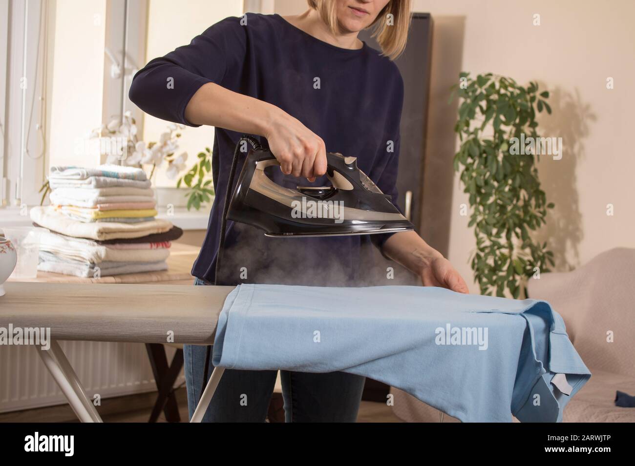 Die Frau hält ein Bügeleisen, aus dem Dampf austritt. Bügelkleidung auf  einem Bügelbrett. Gebügelte Kleidung liegt im Hintergrund auf dem Tisch  Stockfotografie - Alamy