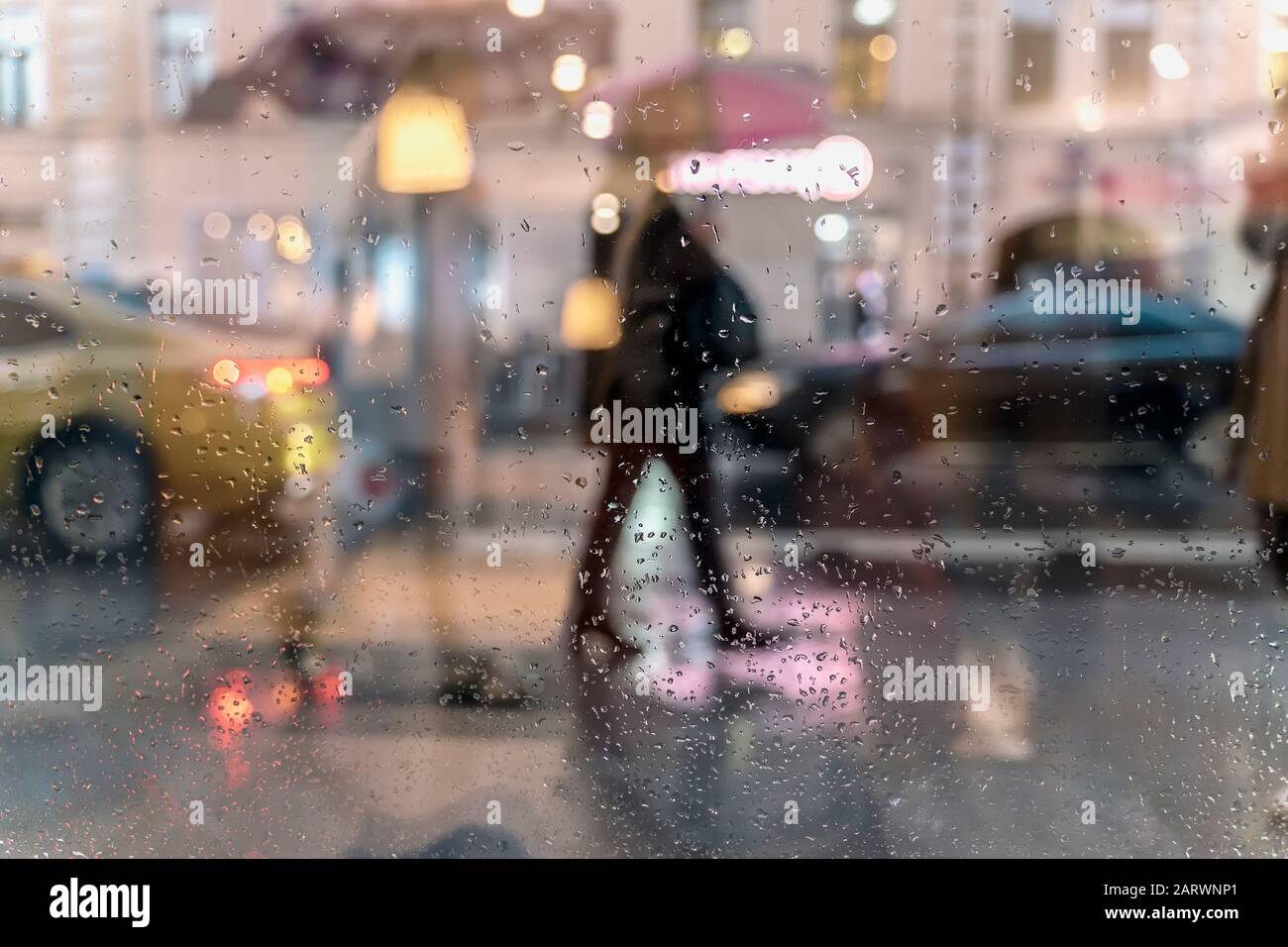 Verschwommene ReflexionsSilhouetten der Menschen beim Wandern bei Regen unter Sonnenschirmen und Bokeh-Stadtlicht, Nacht. Blick durch Glasfenster mit Regen Stockfoto