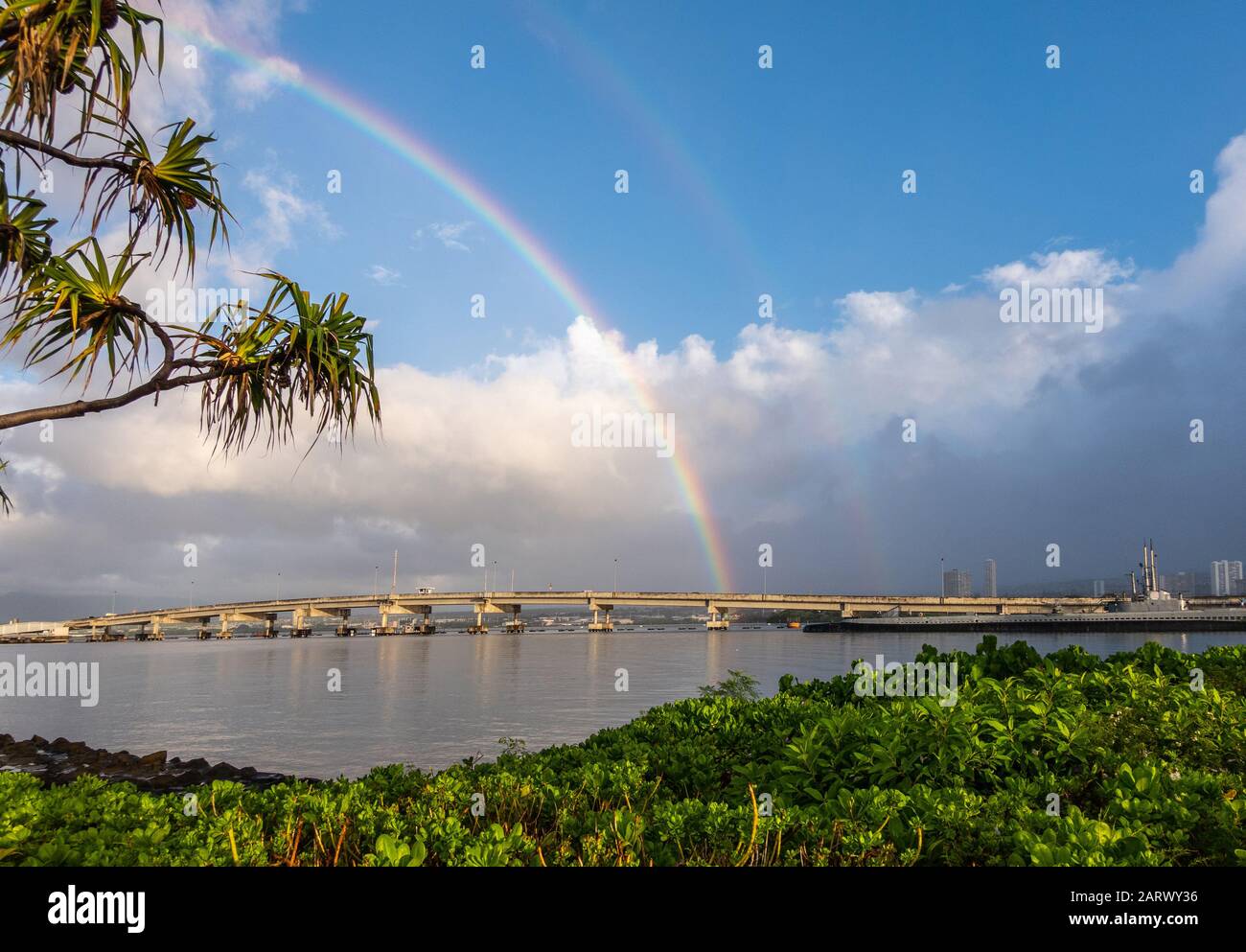 Oahu, Hawaii, USA. - 10. Januar 2020: Pearl Harbor. Ford Island Bridge toubhed by Double Regenbogen under Blue Sky with Heavy Rain Clouds. Grünes Laub Stockfoto