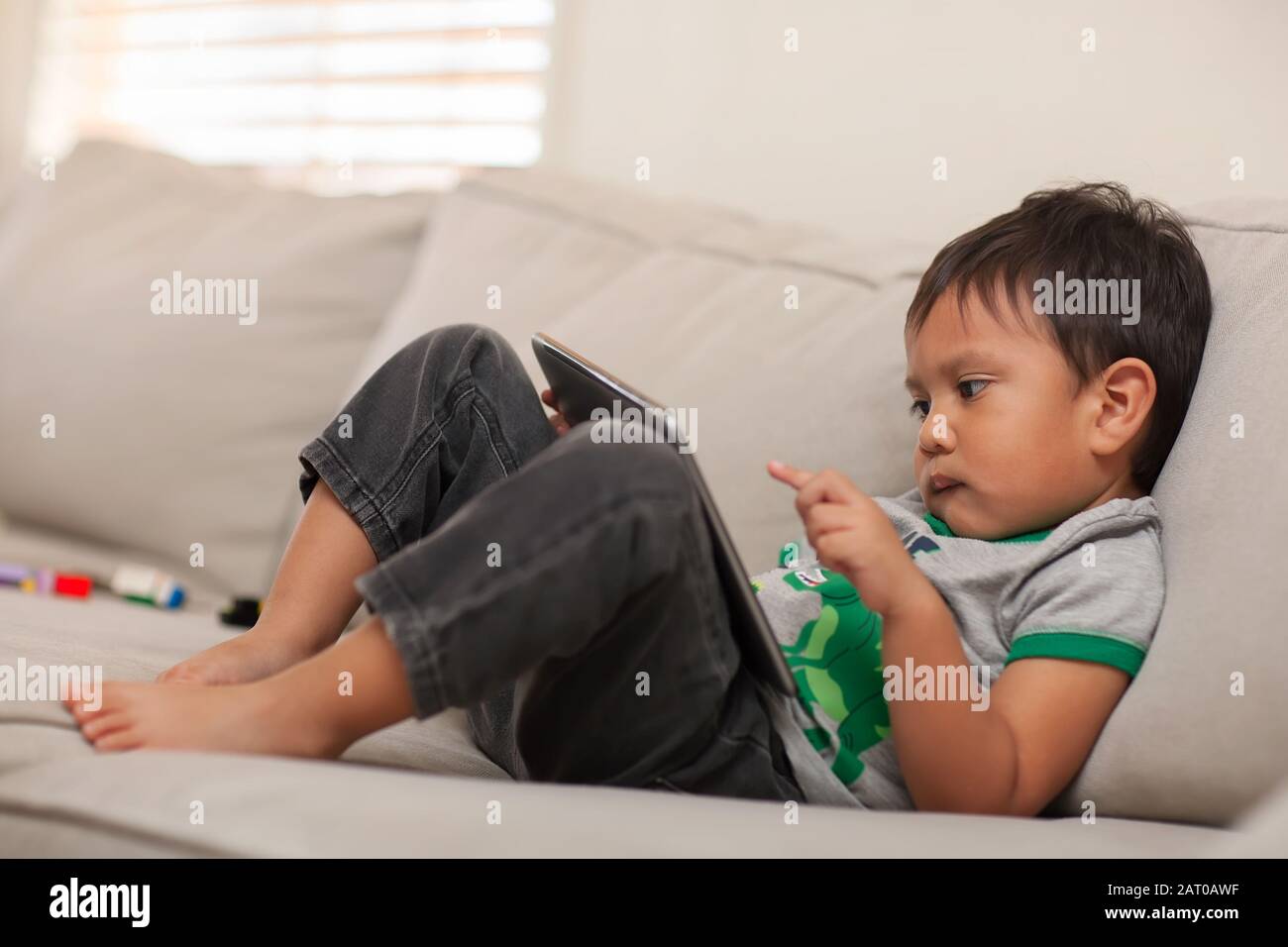 Ein junges Kind, 3 Jahre alt, das seinen kleinen Finger auf dem Touchscreen seines großen Tablets verwendet, während er das Internet auf einer bequemen Couch beobachtet. Stockfoto