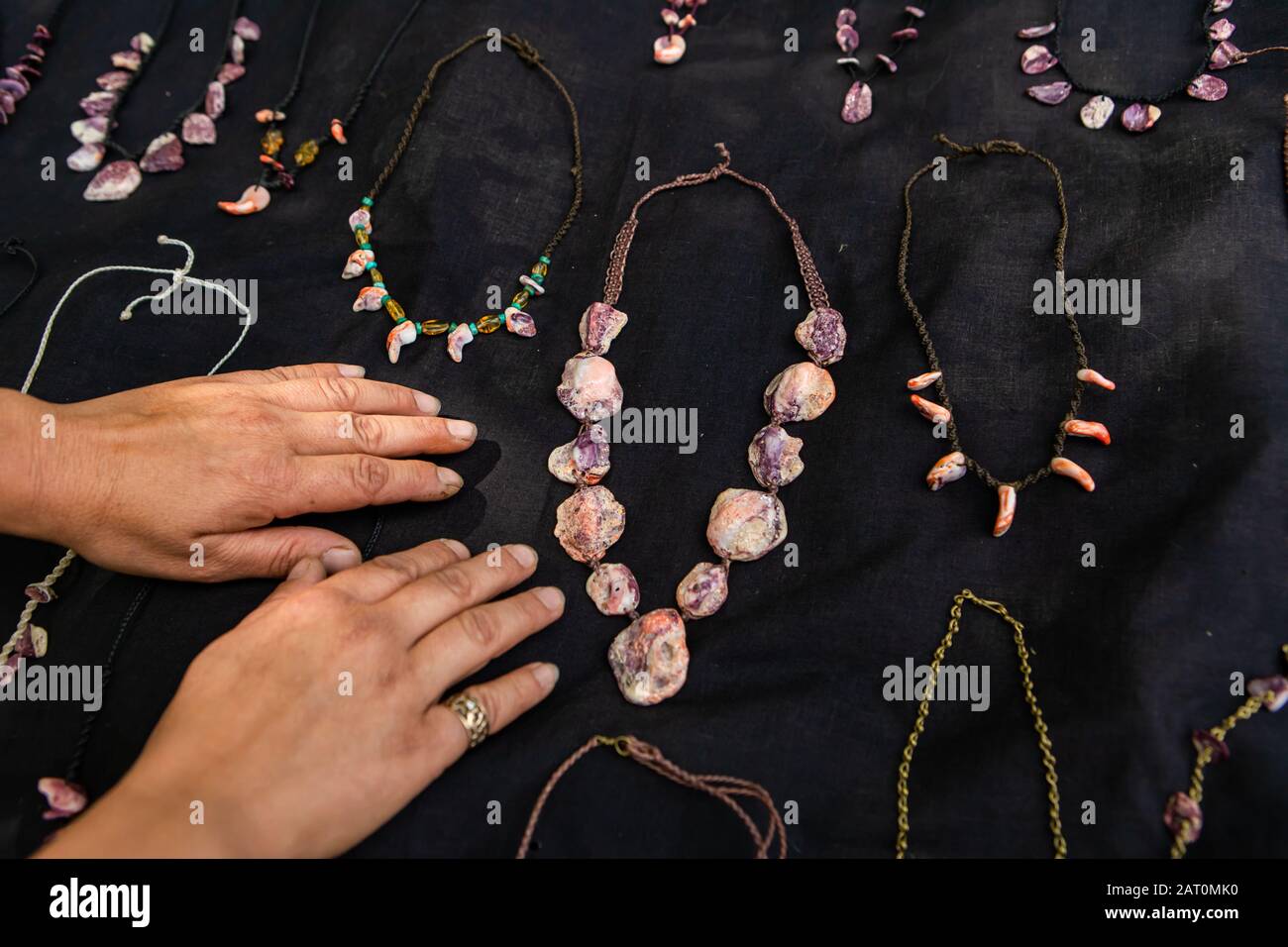 Nahaufnahme der Hände des Mannes, die auf dem schwarzen Samttuch eines Bauernmarkt-Juweliergeschäftes aufliegen. Halsketten aus Edelsteinen und Steinen rundum. Stockfoto