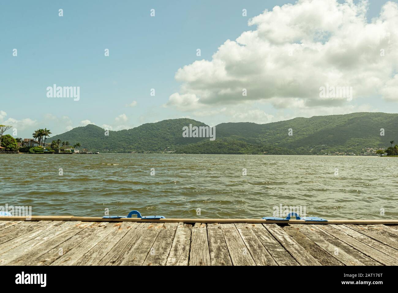 Alter Holzsteg über einem See (Lagoa da Conceição - Florianopolis - Brasilien). See mit einer Welligkeit umgeben von Vegetation und Bergen an einem sonnigen Tag. Stockfoto