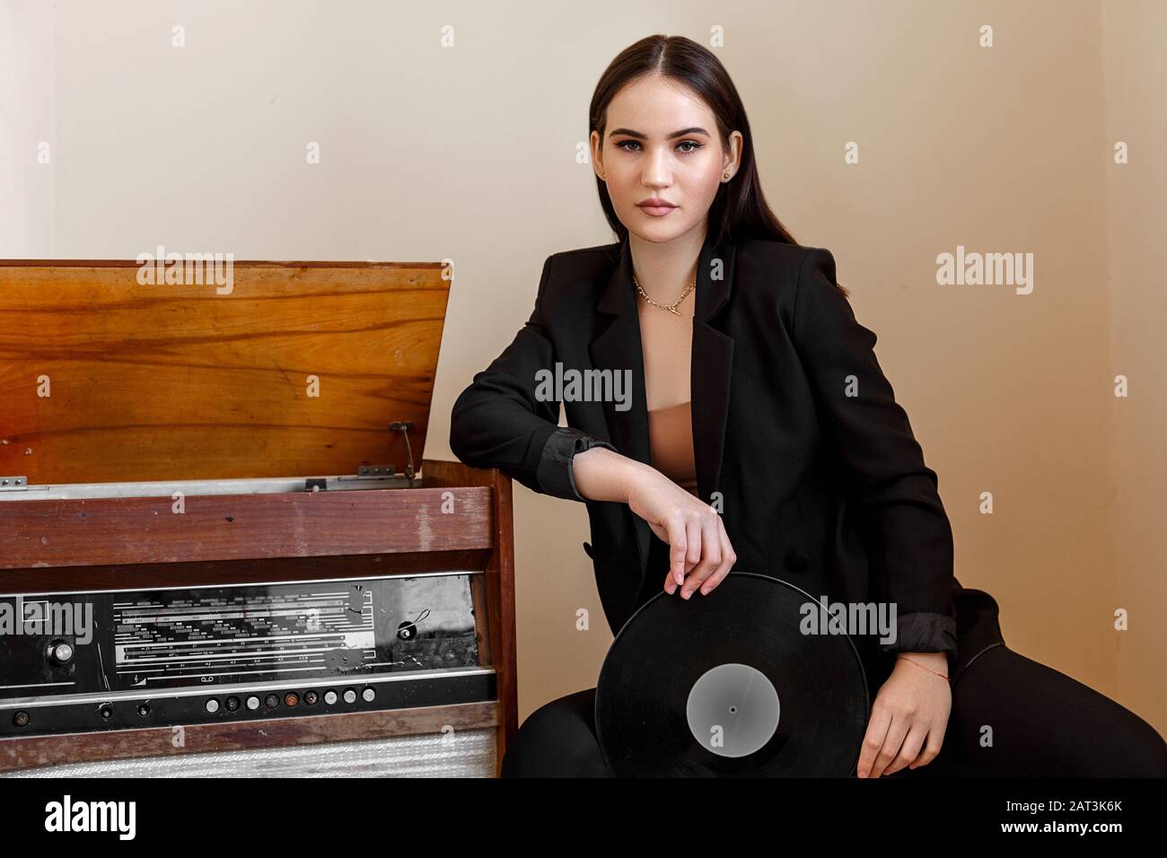 Eine schöne kaukasische junge Frau in einem schwarzen Anzug und schwarzen Sandalen stellt sich neben einem Vintage-Plattenspieler mit einer Grammophon-Schallplatte in ihren Händen Stockfoto