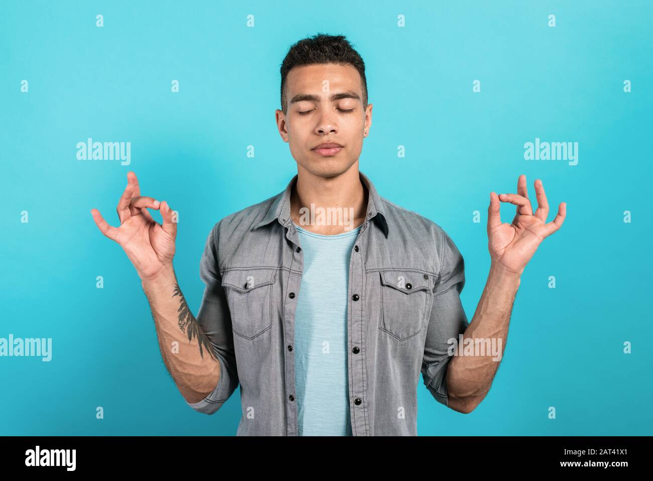 Der Mensch meditiert, während er vor einem blauen Hintergrund steht, seine Augen schließt und seine Arme in Yoga-Mudra hält Stockfoto