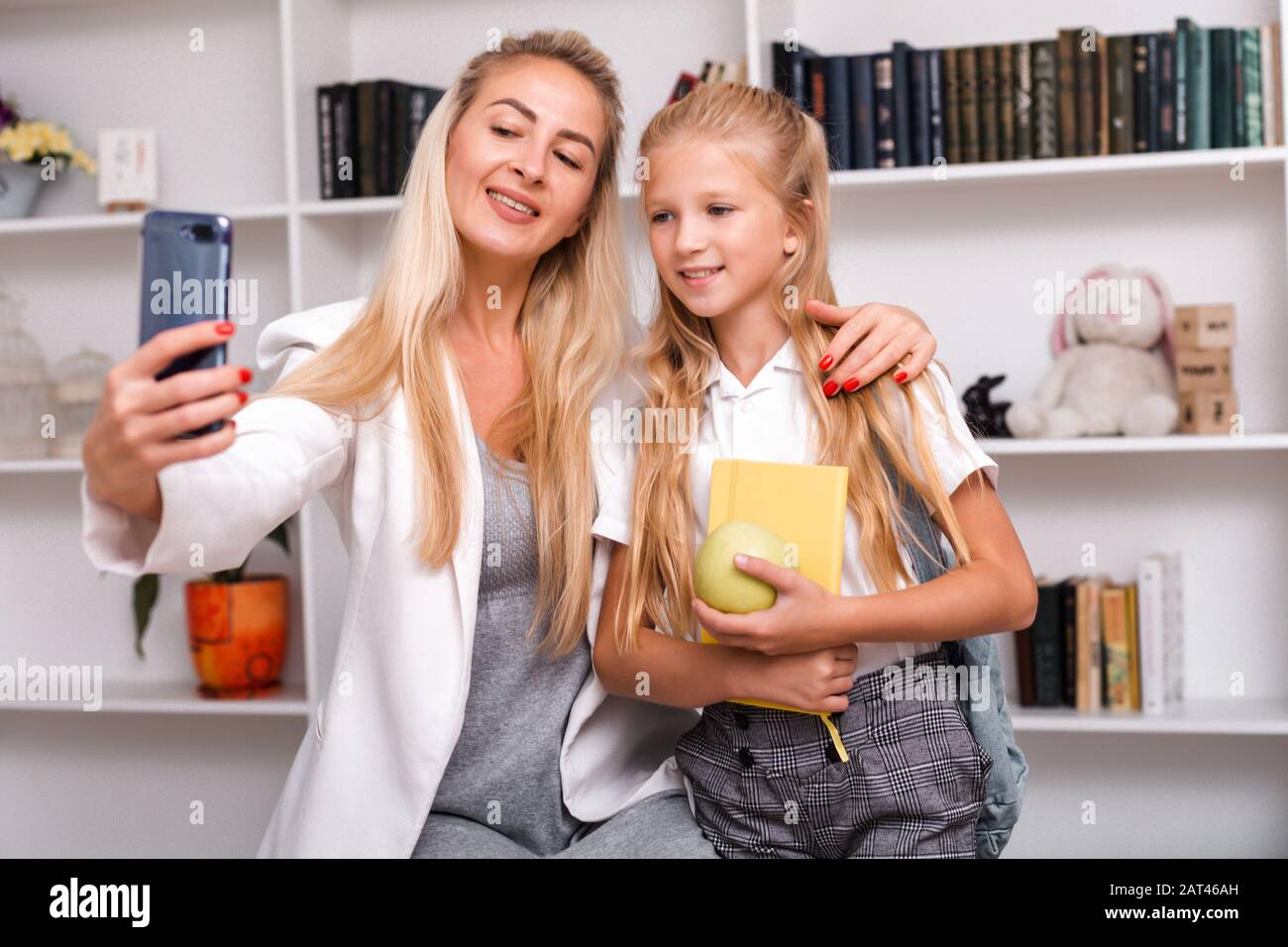 Süße Mutter und kleine Tochter mit Rucksack und Buch in den Händen, die zu Hause Selfies machen Stockfoto