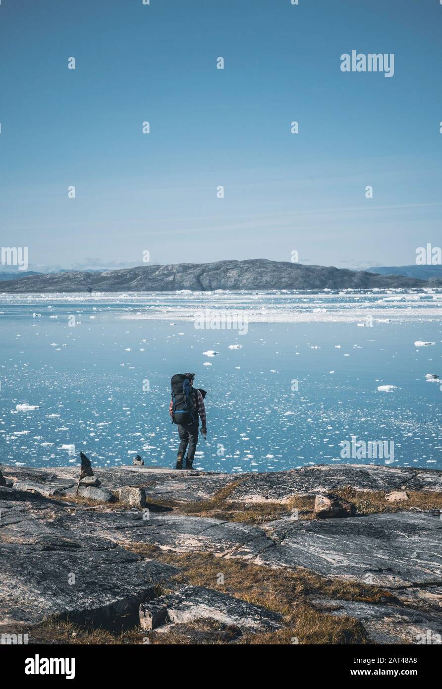 Junger weißer kaukasier mit Rucksack auf einer Wandertour in Grönland mit Eisbergen und Blick auf den Atlantik. Arktische Landschaft im Sommer. In Der Nähe Von Arcti Stockfoto