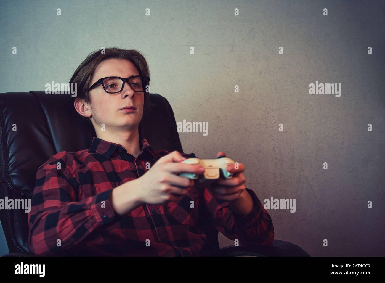 Der ernsthafte Jugendliche Junge, der spät in der Nacht Videospiele spielt, sitzt entspannt in seinem Sessel über grauem Wandhintergrund. Vorsatz Teenager Mann hält einen Joystick Con Stockfoto