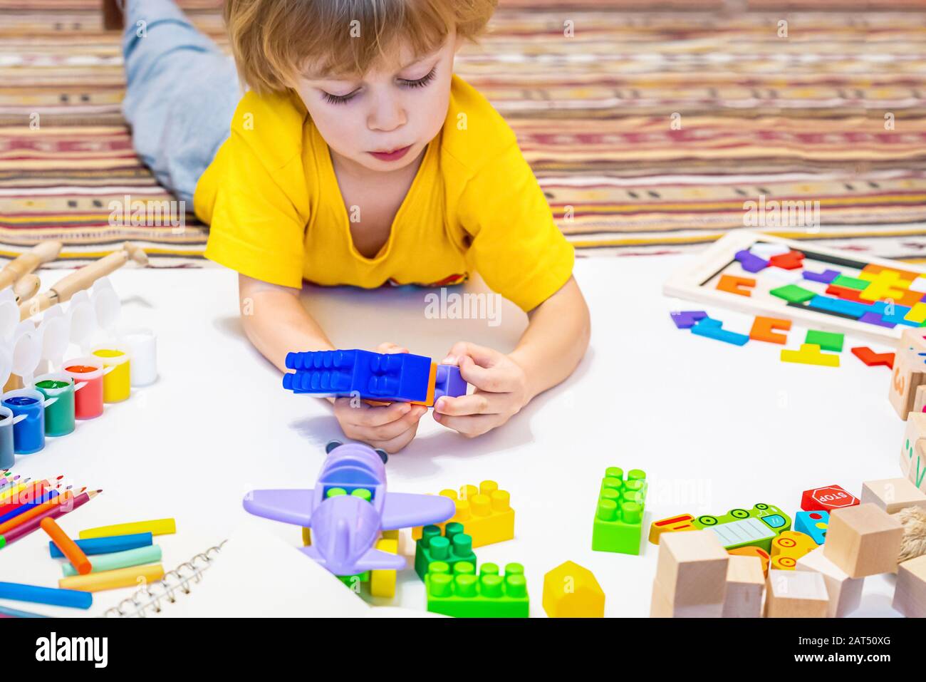 Prescool Junge spielen mit Blöcken, Flugzeugen und Autos. Pädagogisches Spielzeug für Kinder in der Vorschule und im Kindergarten, das auf Papier auf dem Boden steht. Kind zu Hause oder in der Kindertagesstätte. Stockfoto