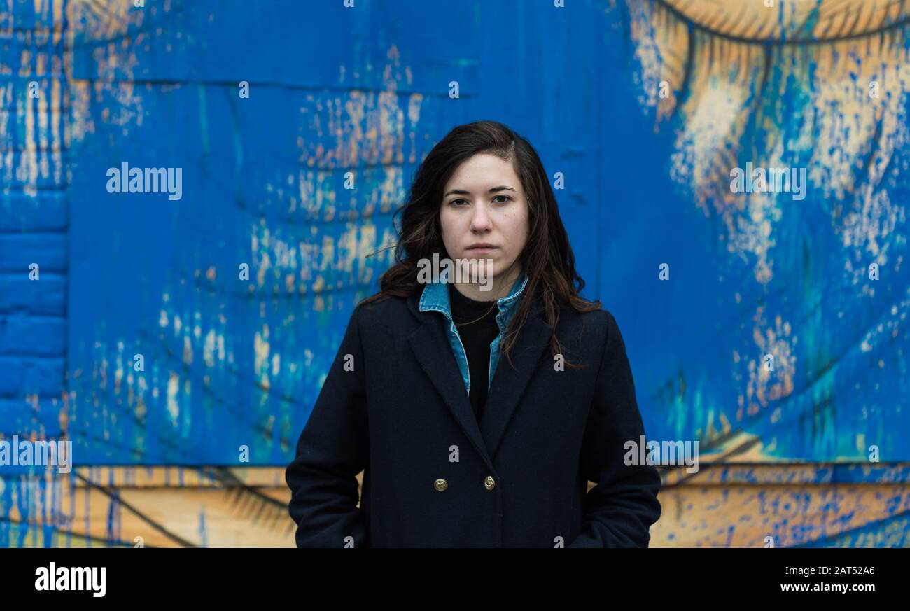 Attraktive junge weiße Frau mit braunen Haaren, die gegen eine blaue Graffitiwand posiert Stockfoto