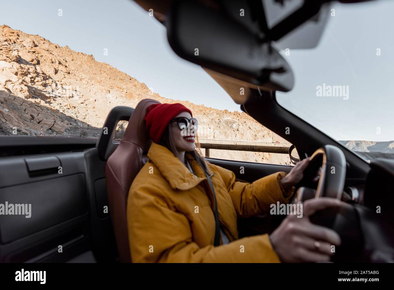 Fröhliche Frau in hellem Hut und Jacke, die mit einem Cabrio-Auto unterwegs ist, während sie bei Sonnenuntergang auf der Wüstenstraße unterwegs ist. Das Bild, das auf den Hintergrund fokussiert ist, ist Frau nicht im Fokus Stockfoto