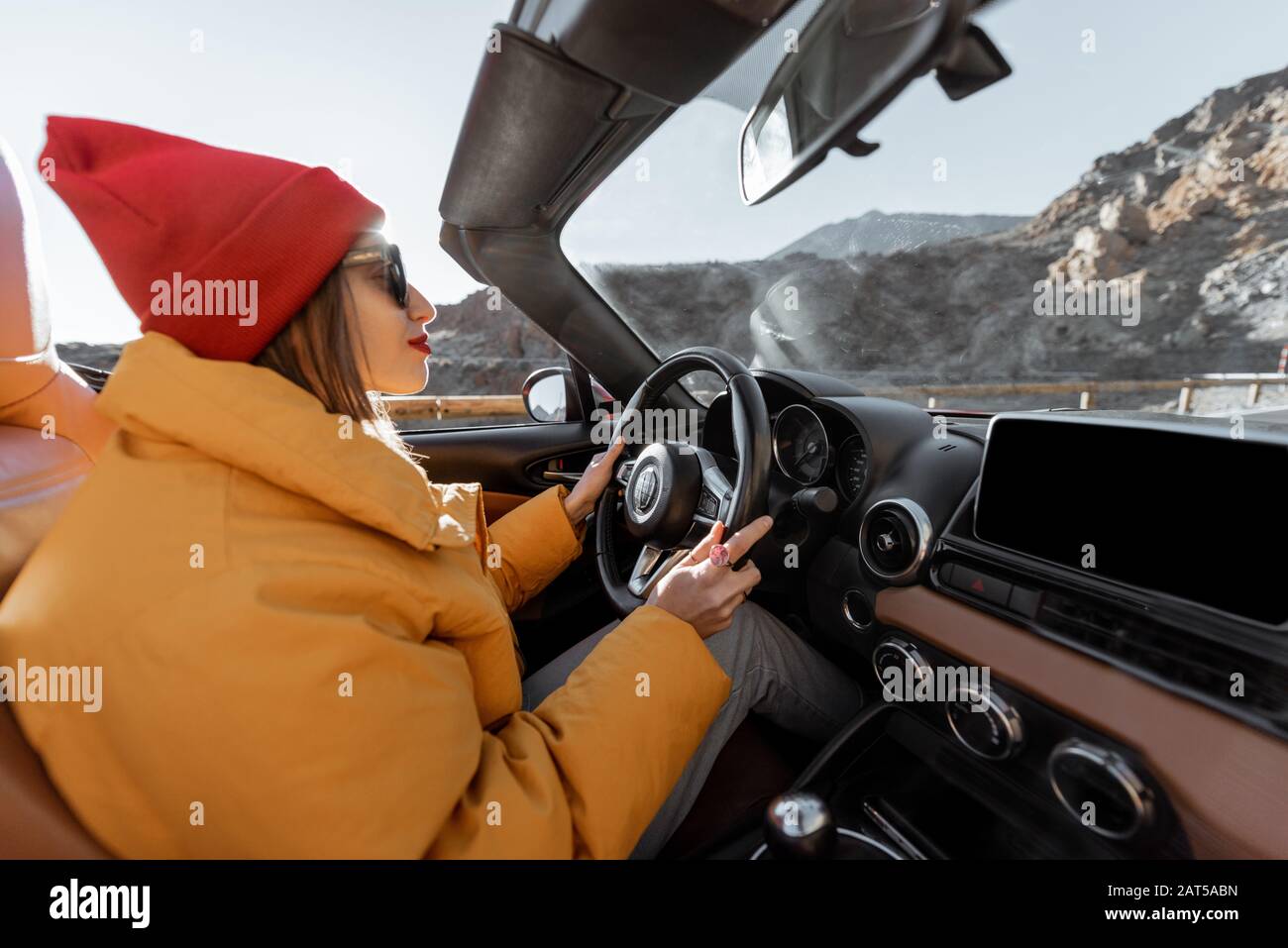 Fröhliche Frau in hellem Hut und Jacke, die mit einem Cabrio-Auto unterwegs ist, während sie bei Sonnenuntergang auf der Wüstenstraße unterwegs ist. Sorgloses Lifestyle- und Reisekonzept Stockfoto