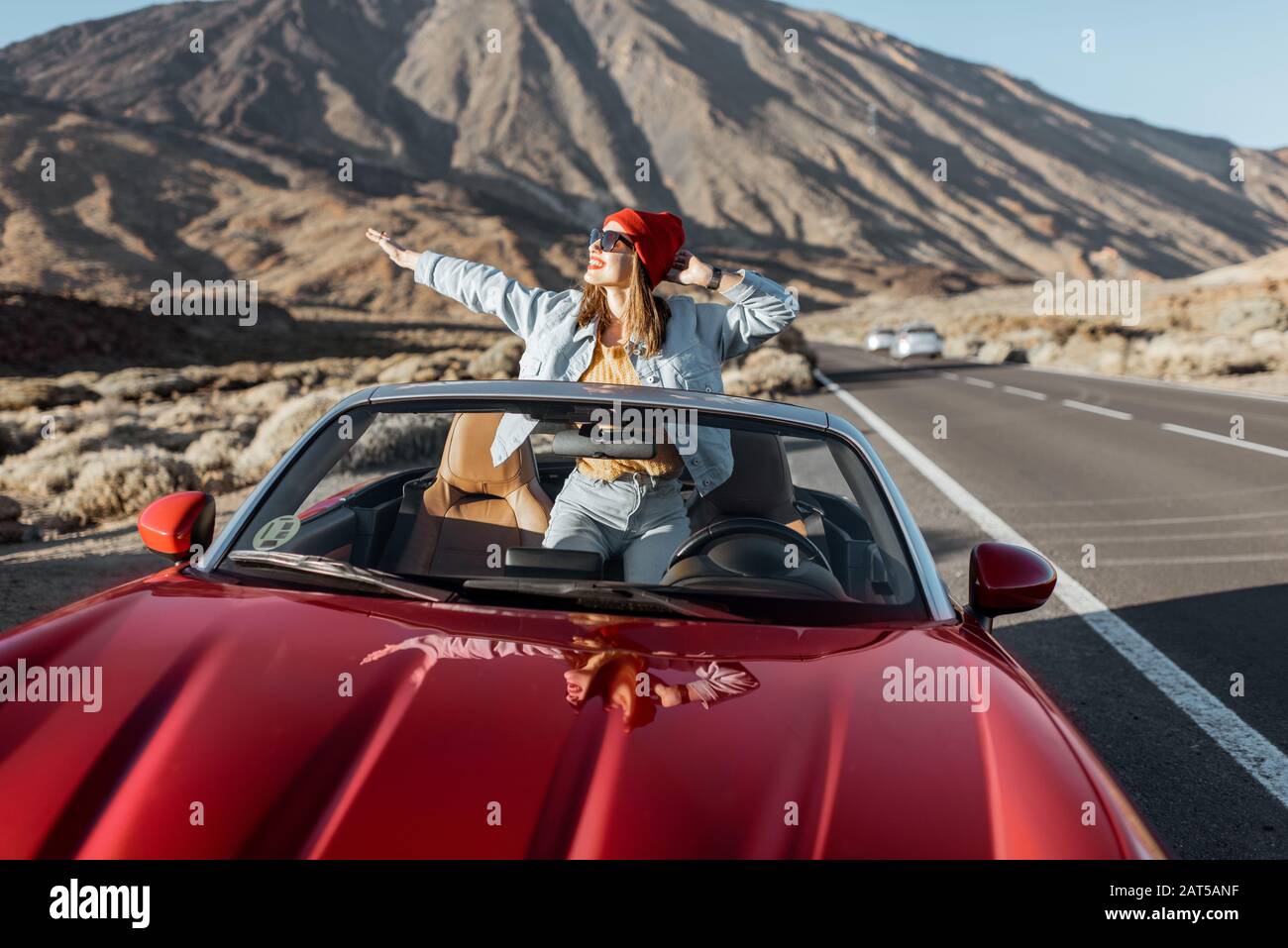 Unbeschwerte Frau, die sich beiläufig in Jeans und rotem Hut kleidete und die Fahrt auf der Straße im vulkanischen Tal genossen und die Hände von einem wandelbaren Auto am Straßenrand hob Stockfoto