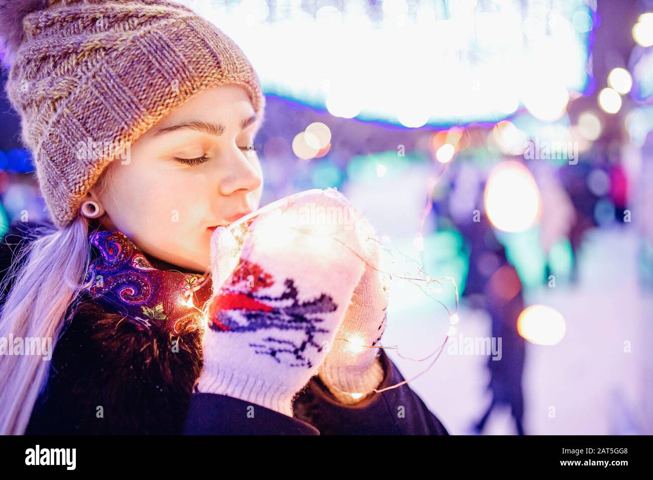 Wunderschönes Mädchen trinkt im Winter heißen Tee aus Pappglas auf dem Hintergrund der abendlichen Bokeh-Beleuchtung Stockfoto