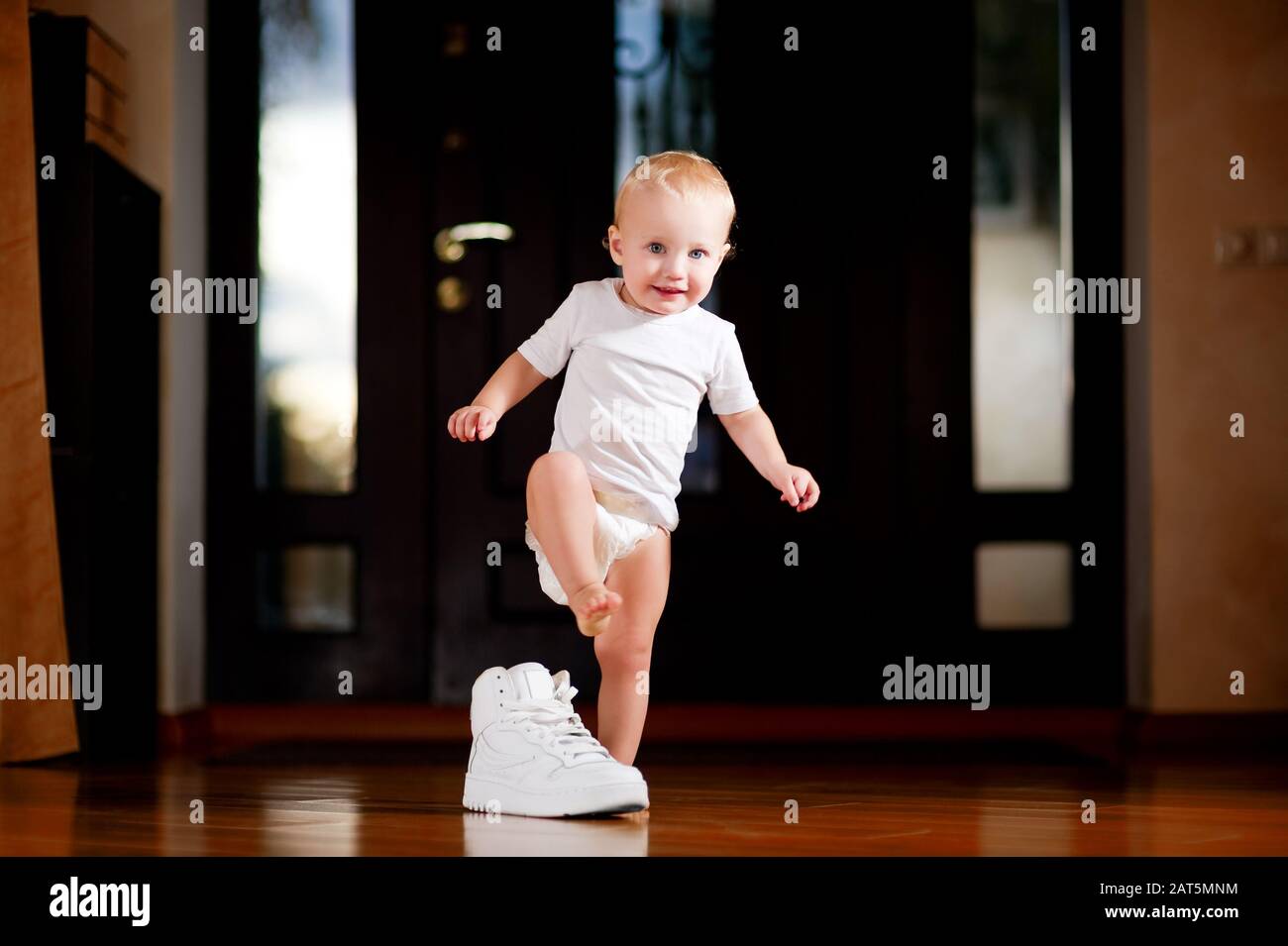 Ein Kind steht neben einem weißen Sneaker für Erwachsene, der im Flur steht und spielerisch lächelt Stockfoto